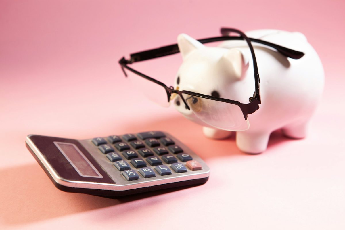 A white piggy bank wearing glasses beside a calculator on a pink background